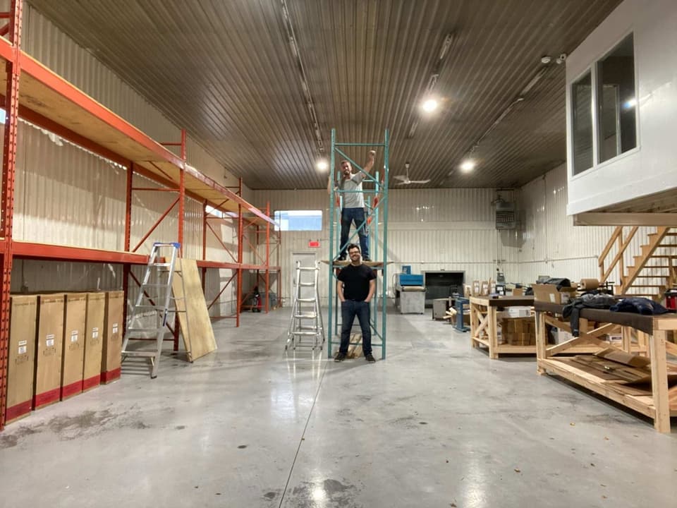 Simon, fist raised on top of a rack, and Jonathan at its base, on the day of a first order milestone; beside them, the homemade packing tables